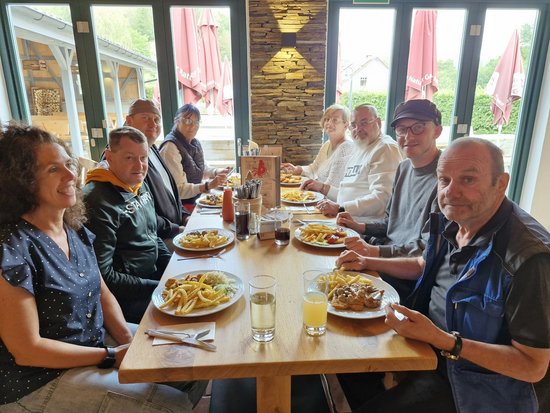 Eine Gruppe von acht Personen sitzt an einem langen Holztisch in einem hellen Restaurant. Vor ihnen stehen Teller mit Pommes frites und Getränken. Im Hintergrund sind große Fenster und geschlossene Sonnenschirme zu sehen. Alle schauen freundlich in die Kamera.