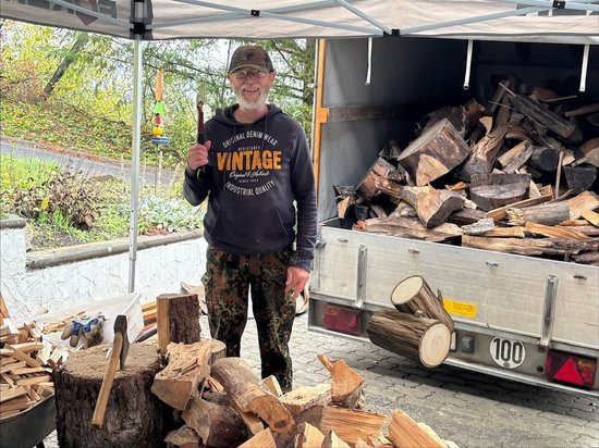 Ein lächelnder Mann steht unter einem Zelt neben einem Anhänger, der voll mit gespaltenem Brennholz ist. Er hält ein Werkzeug in der Hand. Im Vordergrund liegen weitere Holzscheite und ein Baumstumpf mit einer Axt.