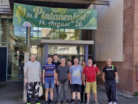 Eine Gruppe von neun Menschen steht vor einem Gebäudeeingang. Darüber hängt ein grünes Banner, das das 34. Platanenfest am 14. August 2026 von 14 bis 18 Uhr im Kulturzentrum Spinnerei ankündigt.