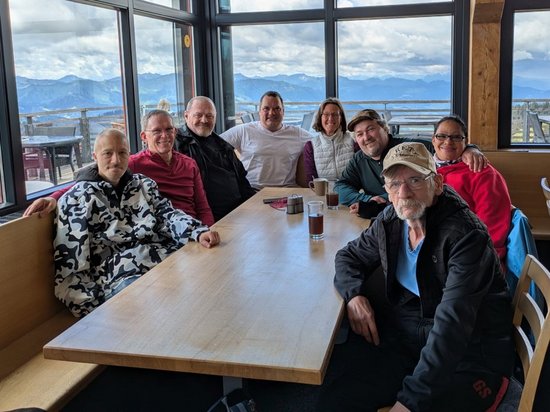 Ein Gruppenfoto von acht Personen, die an einem langen Holztisch in einem Restaurant sitzen. Große Fenster im Hintergrund zeigen eine malerische Berglandschaft unter einem blauen Himmel mit weißen Wolken.